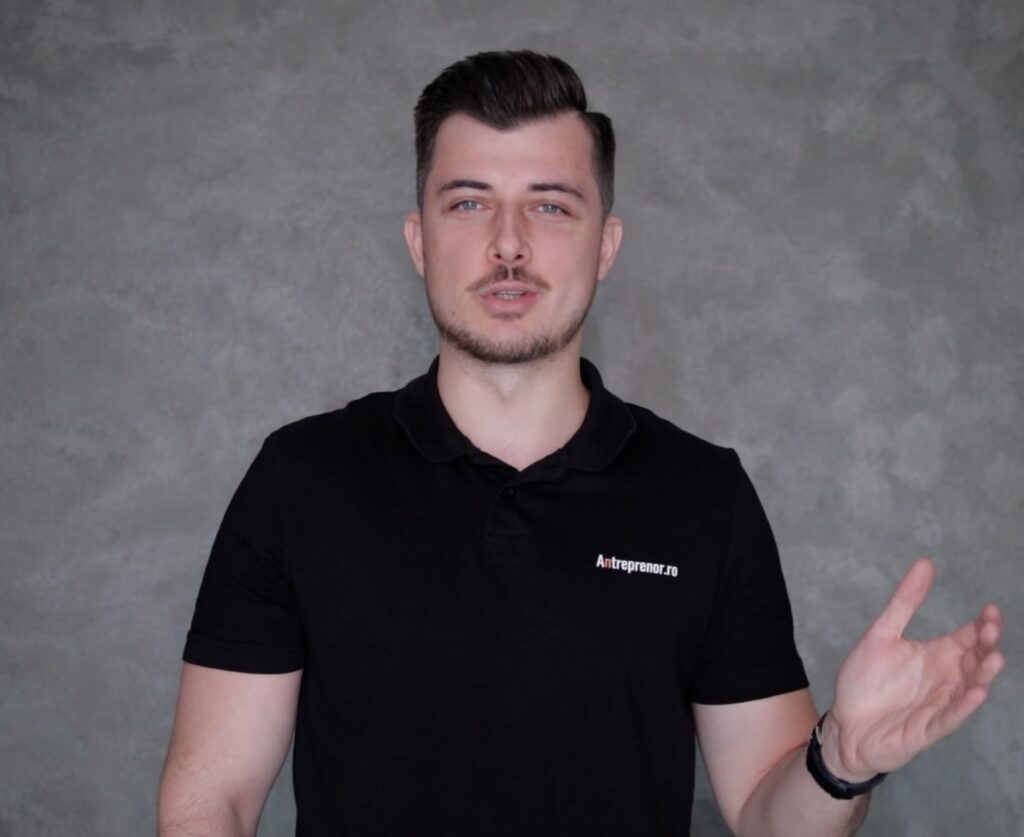 antreprenor.ro- Un bărbat cu părul scurt închis la culoare, purtând un tricou polo negru, stă în fața unui fundal gri texturat, gesticulând cu mâna dreaptă.